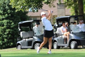 Golfer swings club on lush course, golf carts and spectators in background, sunny day.