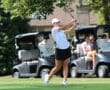 Golfer swings at a golf course with spectators in golf carts watching closely.