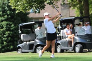 Golfer swings at a golf course with spectators in golf carts watching closely.