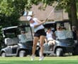 Golfer in a white cap swinging a club on a sunny day with golf carts and spectators in the background.