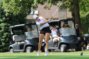 Golfer in a white cap swinging a club on a sunny day with golf carts and spectators in the background.