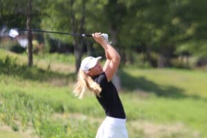 Golfer in black shirt and white cap swinging club on sunny day at lush golf course.