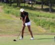 Golfer preparing to swing on a sunny course, wearing a black shirt and white shorts, focused on the ball.