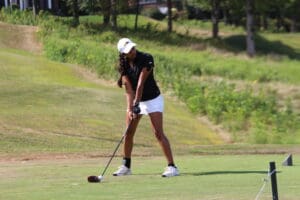 Golfer preparing to swing on a sunny course, wearing a black shirt and white shorts, focused on the ball.