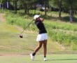 Golfer in mid-swing on a sunny day, wearing a black shirt and white shorts, on a scenic golf course.