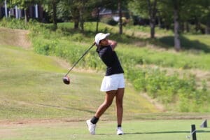 Golfer in mid-swing on a sunny day, wearing a black shirt and white shorts, on a scenic golf course.