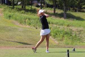 Golfer in action, swinging club on a lush golf course, wearing black top and white skirt.