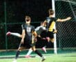 Soccer players in action during night match, one leaping towards goal, others defending.