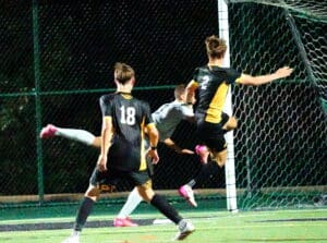 Soccer players in action during night match, one leaping towards goal, others defending.