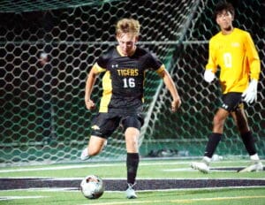 Soccer player in black jersey dribbles towards goal with goalkeeper watching, nighttime match.