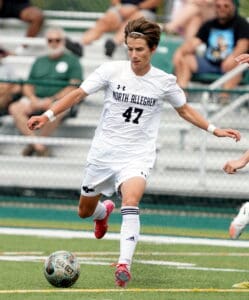 Soccer player in white uniform with North Allegheny dribbles ball on field during a game, fans in background.