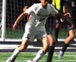 Soccer player in white jersey dribbles ball during nighttime match on grassy field, opponent in black close behind.