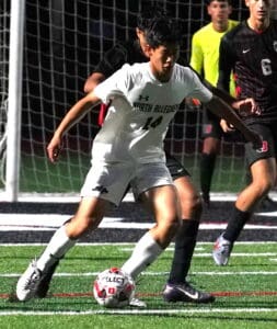 Soccer player in white jersey dribbles ball during nighttime match on grassy field, opponent in black close behind.