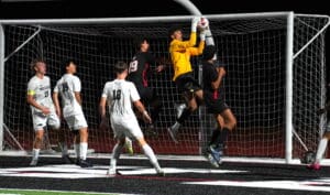 Soccer goalkeeper in yellow saves a shot during a night match with players wearing black and white jerseys.
