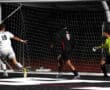 Soccer player in white scores goal past goalkeeper at night game, opposing defender in black approaches.