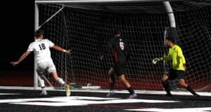Soccer player in white scores goal past goalkeeper at night game, opposing defender in black approaches.