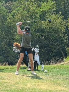 Golfer in mid-swing on grassy course with woods in background, golf bag and clubs nearby.