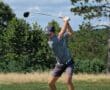 Golfer in mid-swing on a lush, sunny course with trees in the background.