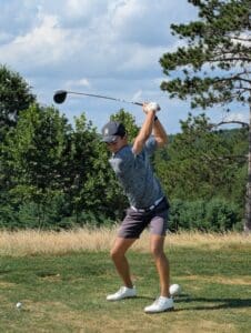 Golfer in mid-swing on a lush, sunny course with trees in the background.