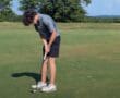 Golfer putting on green under clear sky with trees in background.