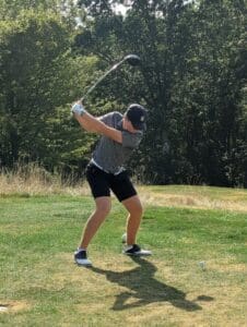 Golfer in mid-swing on a sunny day, surrounded by lush greenery and trees on a golf course.