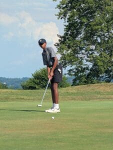 Teen golfer putting on a sunny day at the golf course, surrounded by trees and a clear sky.