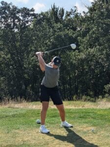 Golfer swinging club at tee on a sunny course, surrounded by lush trees.