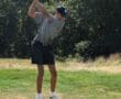 Golfer in mid-swing on a sunny day at the golf course, surrounded by lush green trees.