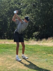 Golfer in mid-swing on a sunny day at the golf course, surrounded by lush green trees.