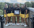 Group of young women in matching outfits holding a first place certificate on a terrace with a scenic backdrop.