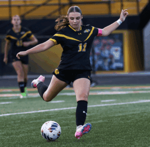 Soccer player in black kit kicking a ball on the field during a match, focused and in motion.