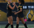 Two female soccer players in black uniforms celebrating on the field with a high five.