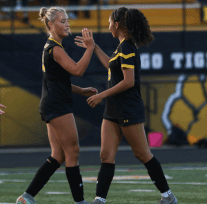 Two female soccer players in black uniforms celebrating on the field with a high five.