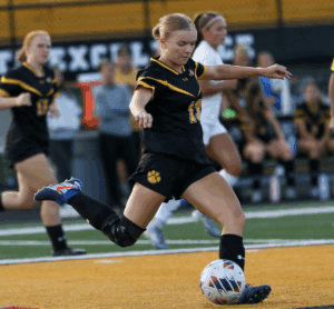 Soccer player in action, kicking the ball with teammates on the field during a match.
