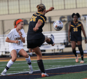 Girls' soccer match action shot with player kicking ball, opponents ready to intercept on a football field.