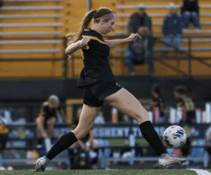 Soccer player in action, kicking a ball during a match on an outdoor field with spectators in the background.