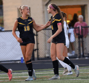 Two female soccer players in black jerseys cheer on the field during a match, showing teamwork and camaraderie.