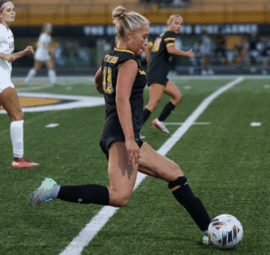 Soccer player in black and yellow uniform kicks ball during a game on a green field.