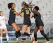 Three female soccer players in black jerseys celebrate on the field during a match, showing team spirit and excitement.