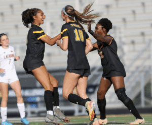Three female soccer players in black jerseys celebrate on the field during a match, showing team spirit and excitement.
