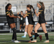 Three female soccer players in black uniforms celebrate on a field after a successful play.