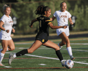 Soccer player in black uniform running with the ball on field, pursued by two opponents in white.