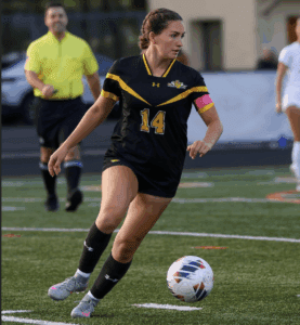 Female soccer player in black jersey dribbles ball on field during match, with referee and opponents in background.