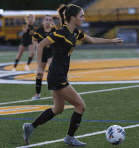 Soccer player in black and yellow uniform dribbling a ball on a green field during a game.