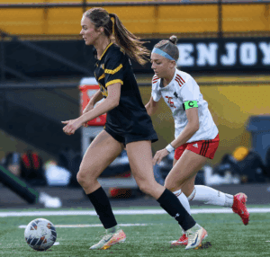 Two female soccer players in action on the field, one wearing black, the other in a white jersey with a captain's armband.