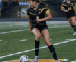 Female soccer player in black and yellow uniform dribbling ball on field during a match.
