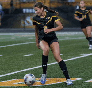 Female soccer player in black and yellow uniform dribbling ball on field during a match.