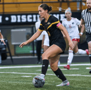 Soccer player in black jersey dribbles ball during match on green field, with opposing team in background.