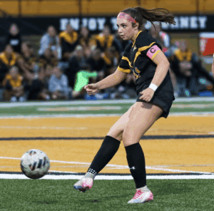 Soccer player in action with pink armband, focusing on kicking the ball during a match on a green field.
