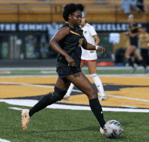 Football player dribbles soccer ball on field during match, wearing black jersey and knee brace.